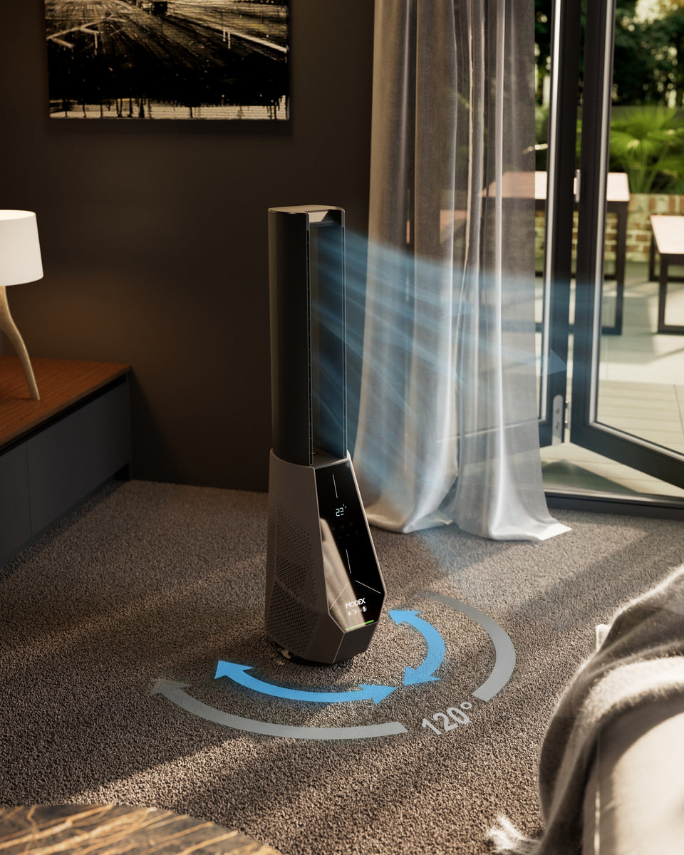 A modern floor-standing tower fan in a dark gray finish, shown on a carpeted floor in a contemporary room with a glass patio door, sheer curtains, and blue graphic overlays illustrating airflow direction and coverage.